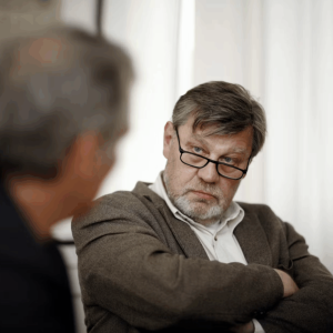Two men sit and talk. One wears a brown sweater and glasses. The other a black coat with his back to the camera.